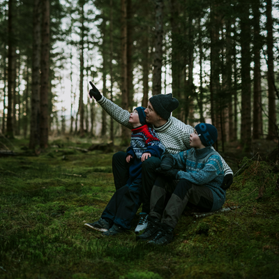 Far med 2 barn på fanget, sitter i skogen og peker mot noe spennende som oppe i trærne