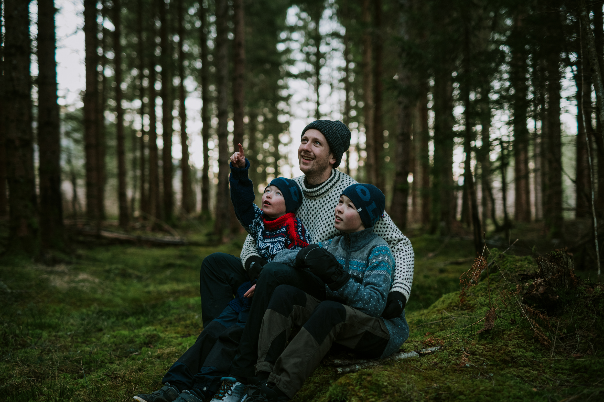 Far med 2 barn på fanget, sitter i skogen og peker mot noe oppe i trærne