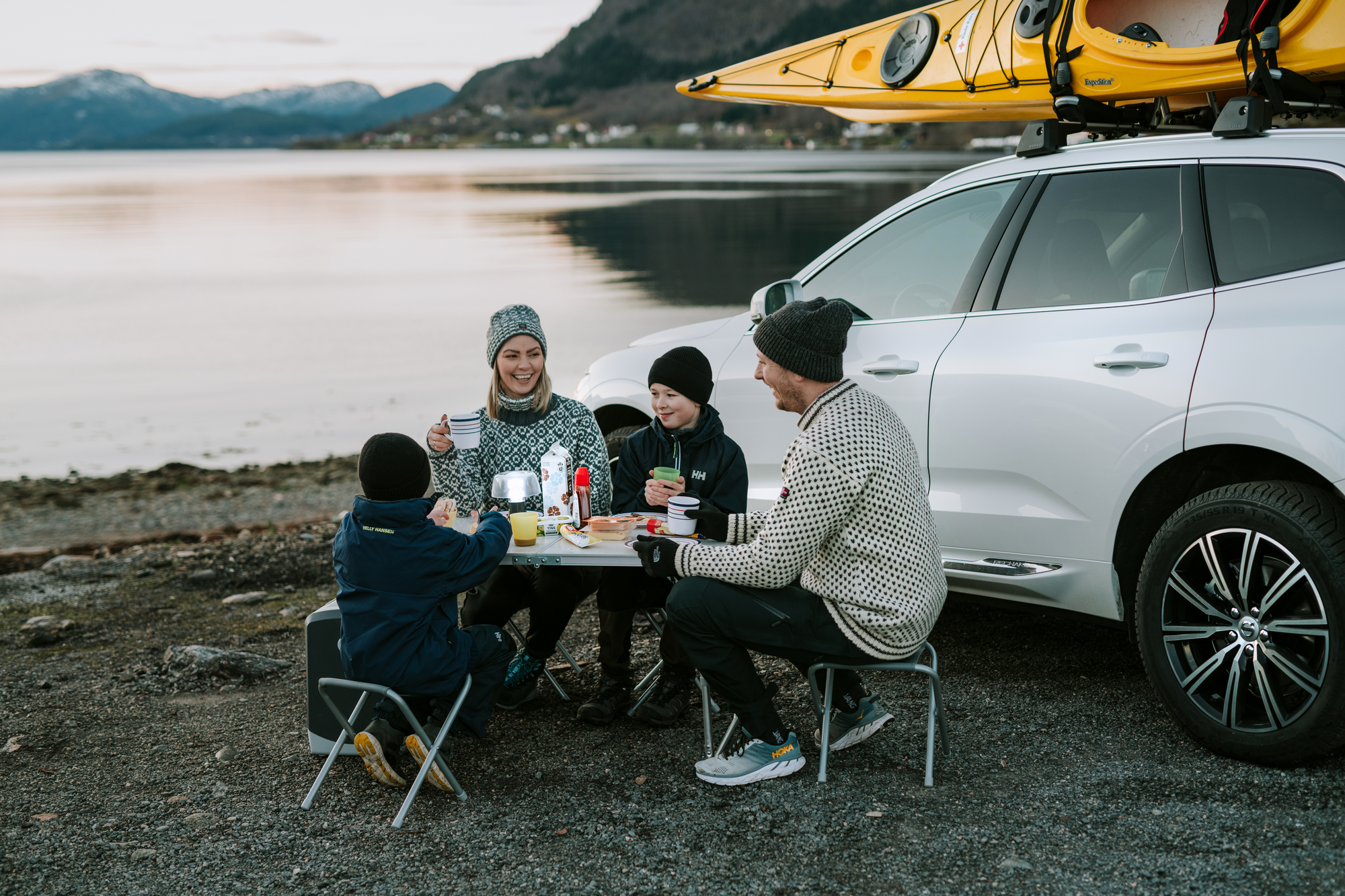 Familie sitter rundt et lite campingbord på stranden, mens de spiser og drikker